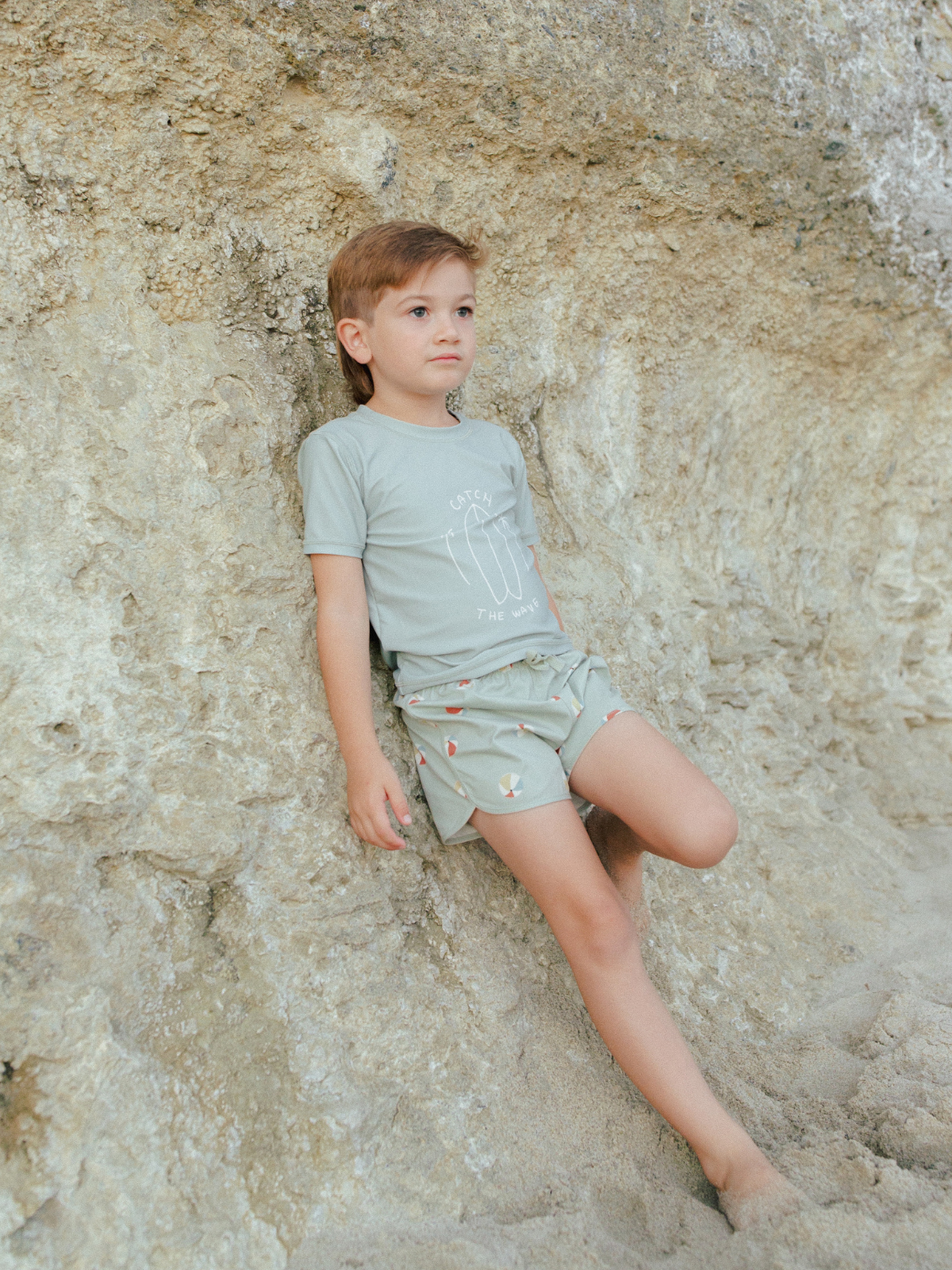 Child in a green outfit poses against a rocky background at the beach, sand visible at his feet.