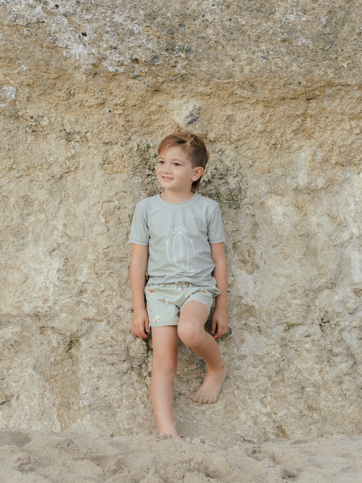 Smiling child in casual beachwear leaning against a rocky wall, with sand beneath and a joyful expression.