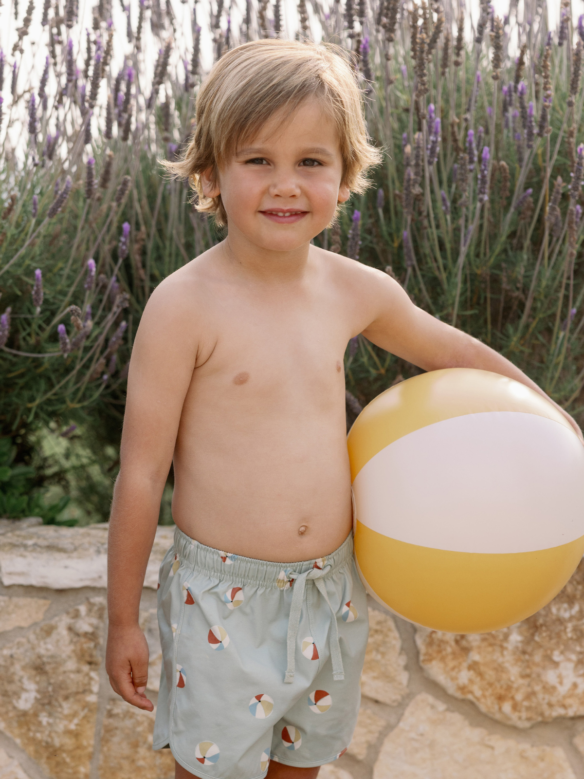 Child in beach shorts holding a beach ball, standing near lavender plants in a sunny outdoor setting.