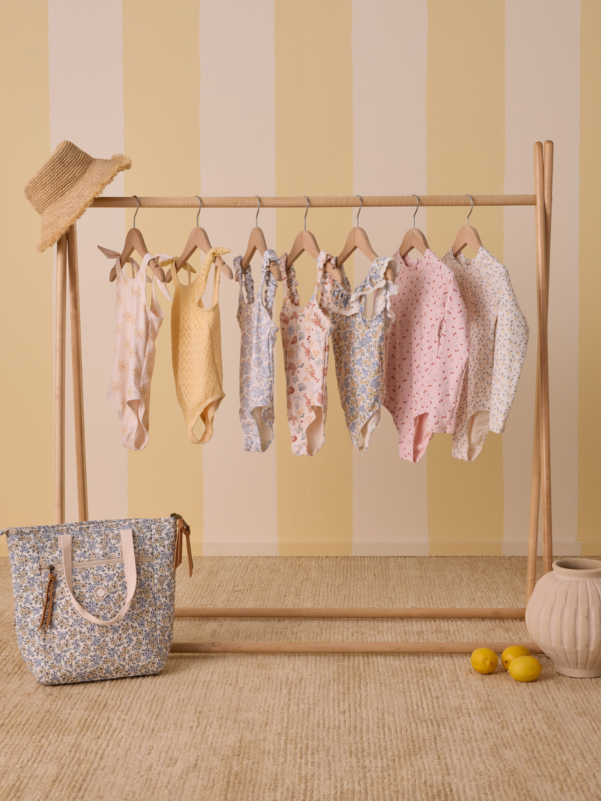 Colorful children's swimsuits hanging on a wooden rack, with a tote bag and lemons on the floor.