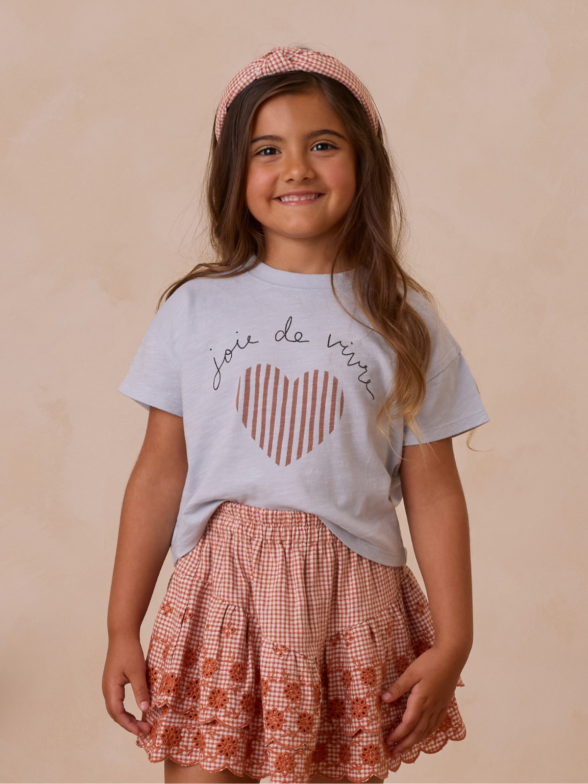 Smiling girl in a light blue shirt with a heart design and a patterned skirt, wearing a matching headband.
