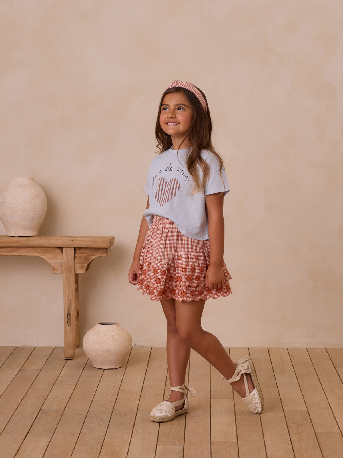 Smiling girl in a heart graphic tee and patterned skirt, standing in a softly decorated room.