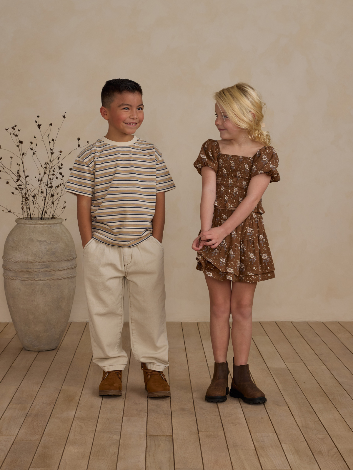 Two children smiling: a boy in a striped shirt and a girl in a floral dress, standing on wooden flooring.