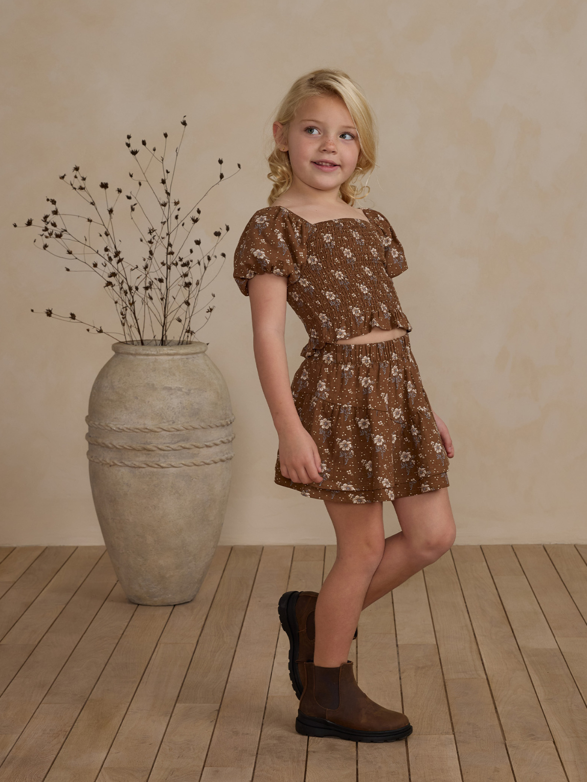 Girl in a floral brown two-piece outfit stands beside a vase against a neutral background.
