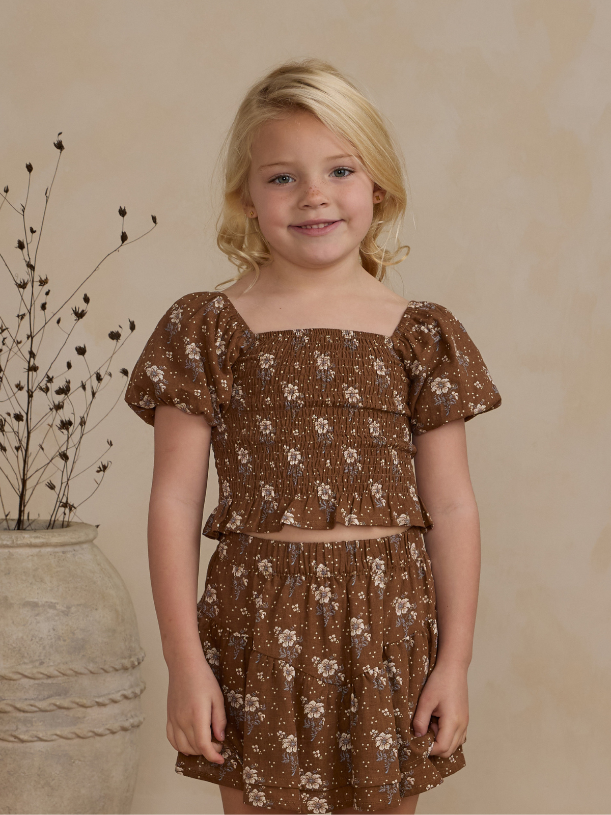 Smiling girl in floral brown outfit, standing against a neutral background with a decorative vase and dried flowers.