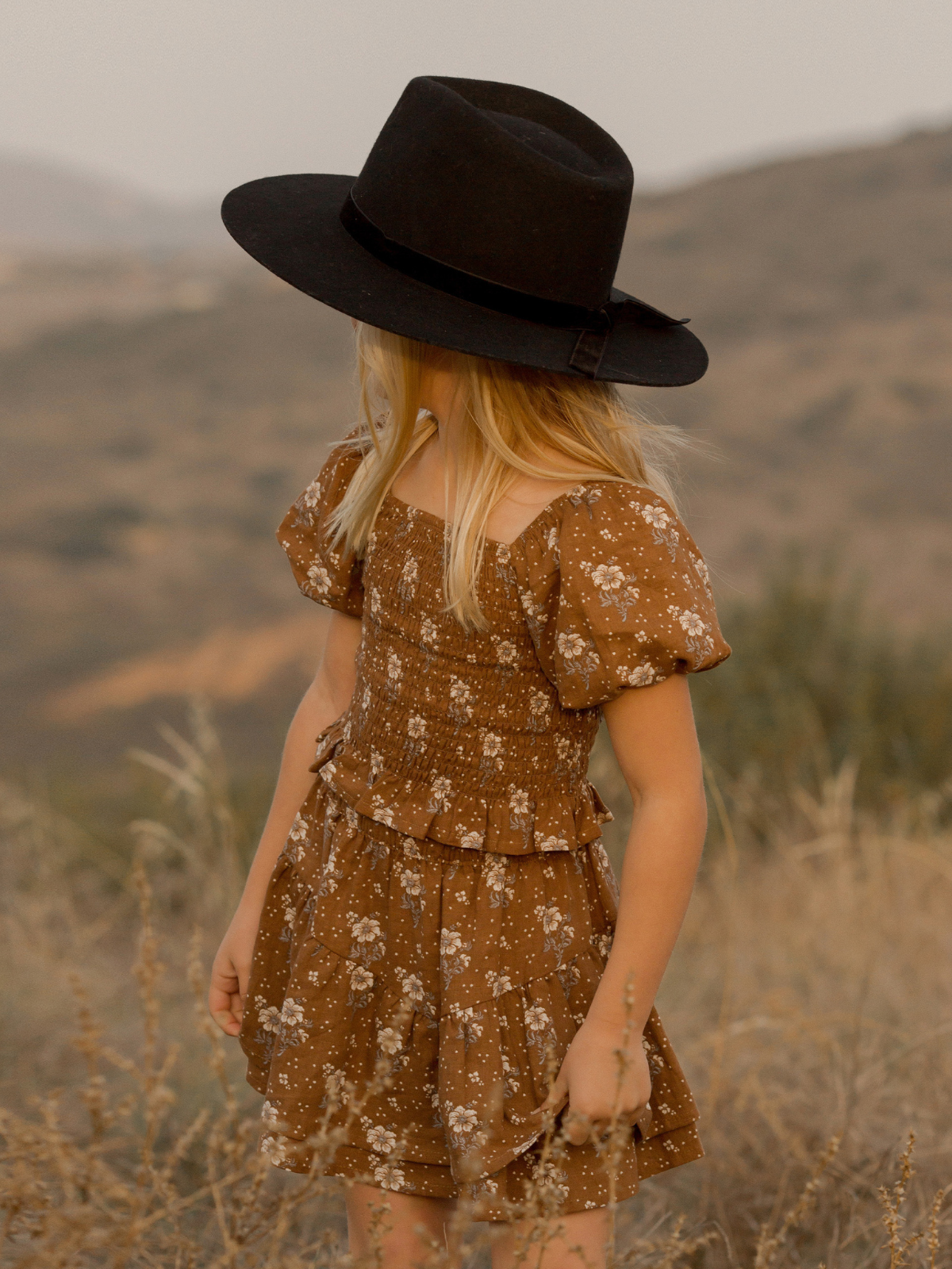 Child in a brown floral dress and black hat stands in a grassy field with mountains in the background.