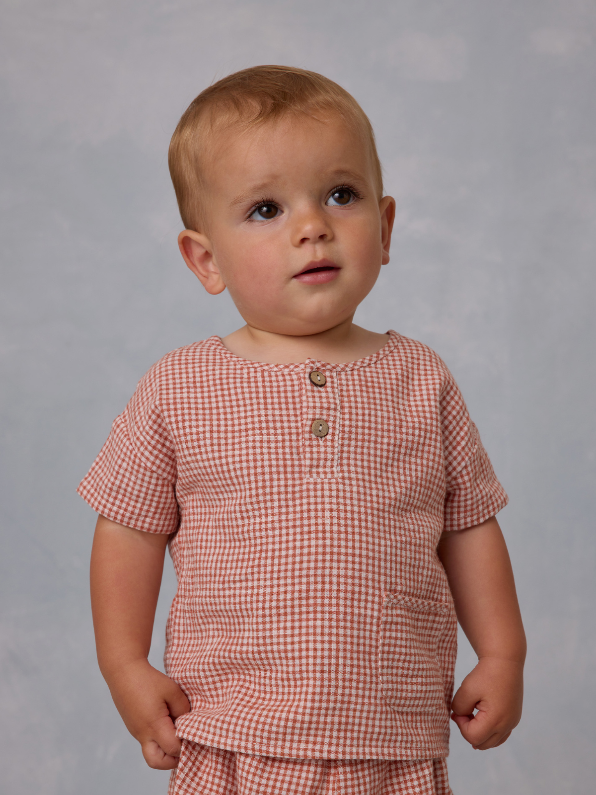 Toddler wearing a short-sleeve checkered shirt, looking curiously at the camera. Light background.