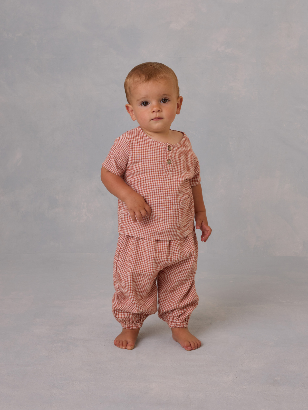 Toddler standing in a checkered outfit with an expression of curiosity against a soft, textured background.