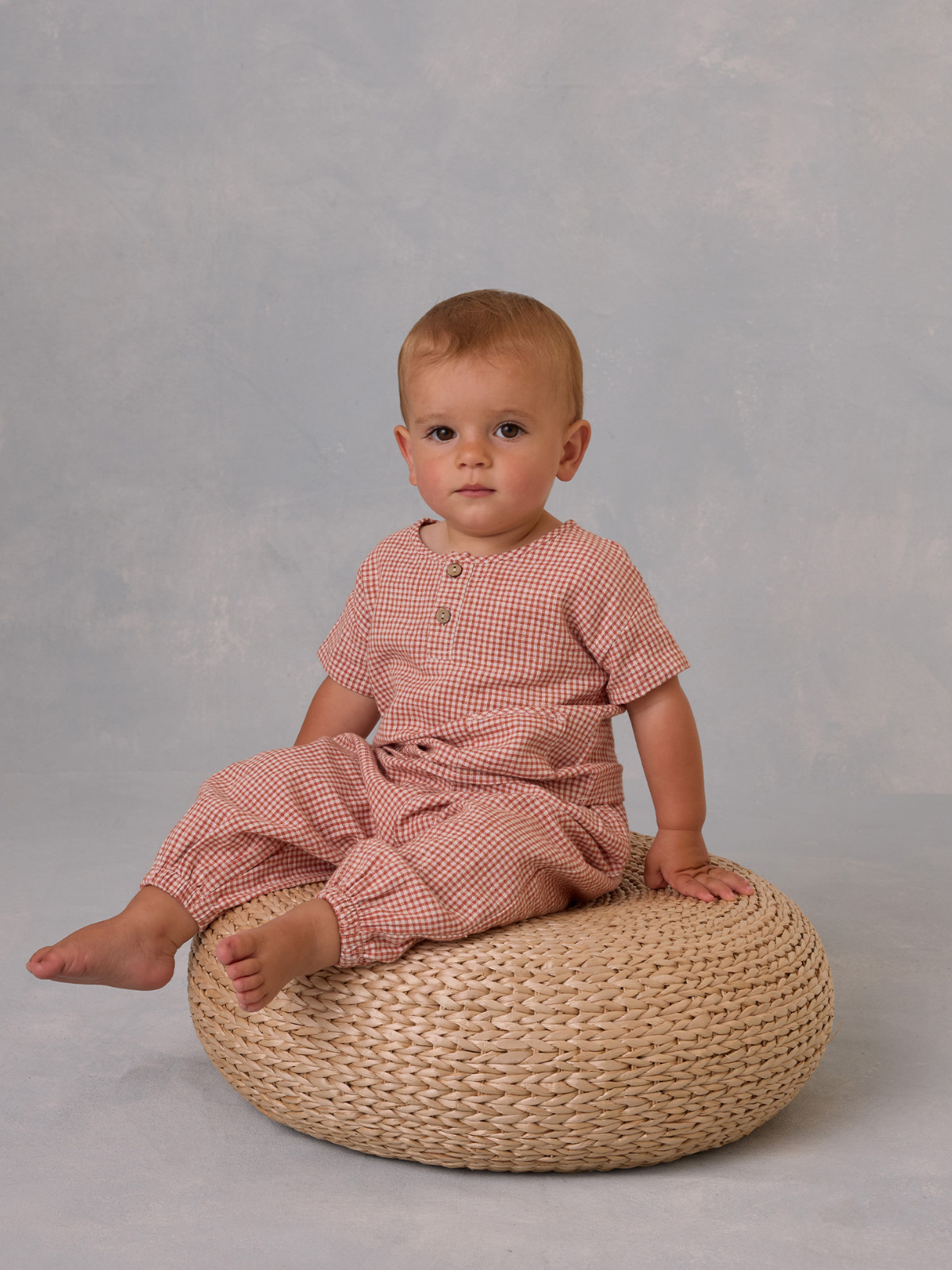 Toddler in a checkered outfit sitting on a woven pouf against a soft blue background.