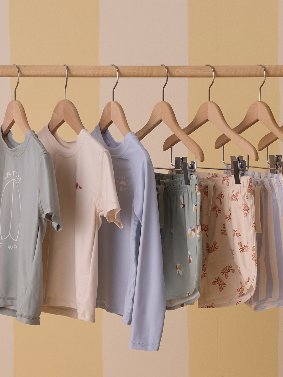 Colorful kids' clothing displayed on wooden hangers against a soft yellow background.