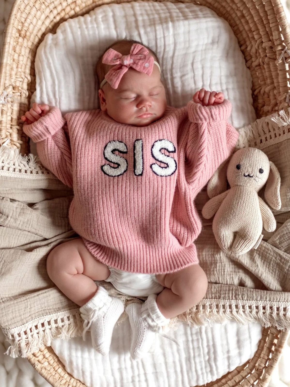 Baby girl in a pink "SIS" sweater sleeps in a basket, with a soft bunny toy beside her. Cozy and peaceful setting.