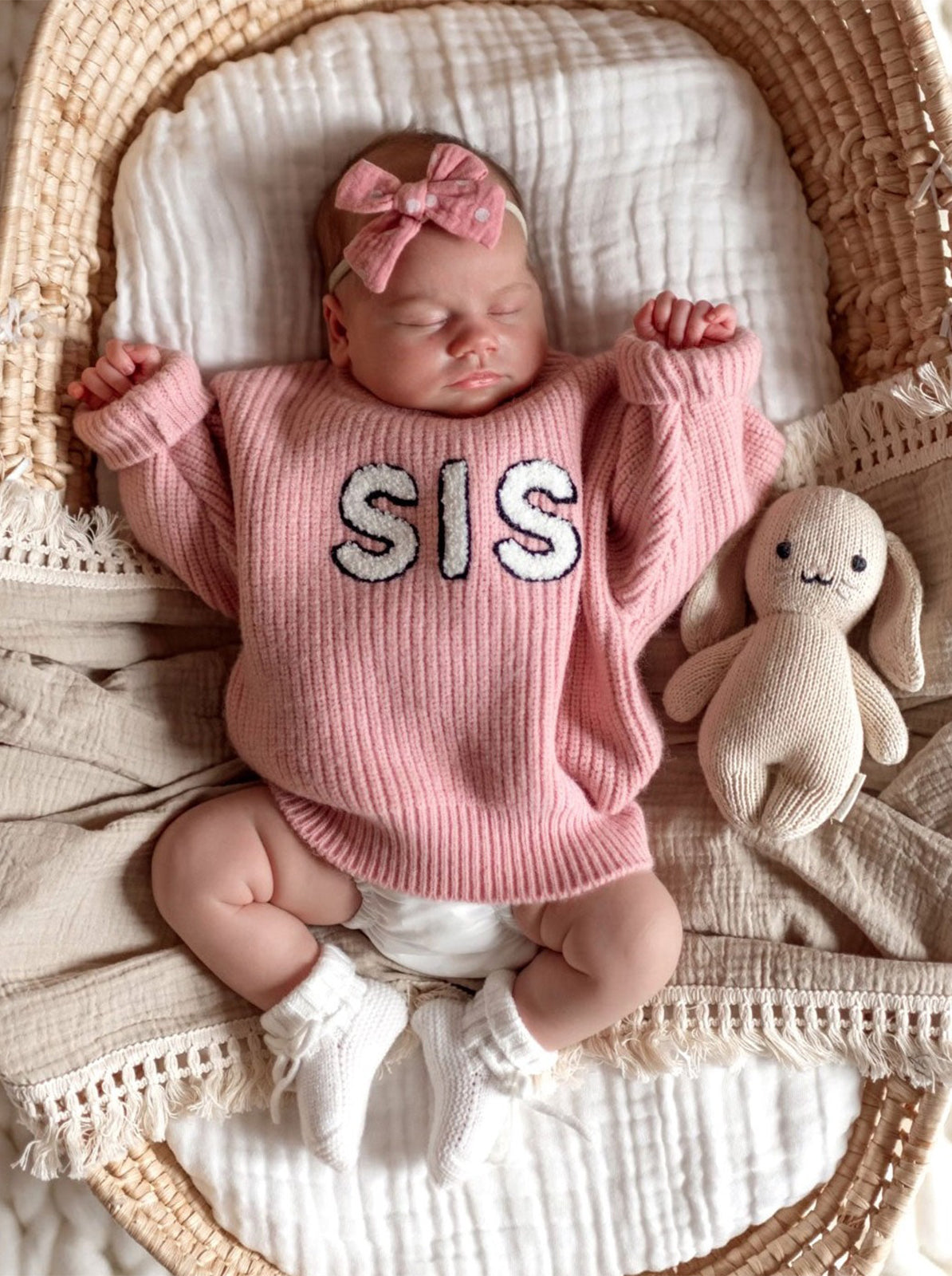 Baby girl in a pink "SIS" sweater sleeps in a basket, with a soft bunny toy beside her. Cozy and peaceful setting.