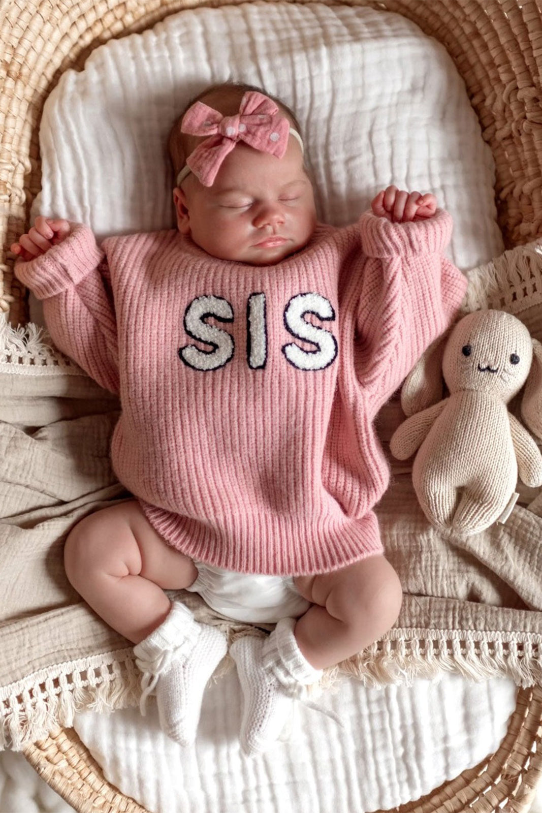 Baby girl in a pink "SIS" sweater sleeps in a basket, with a soft bunny toy beside her. Cozy and peaceful setting.