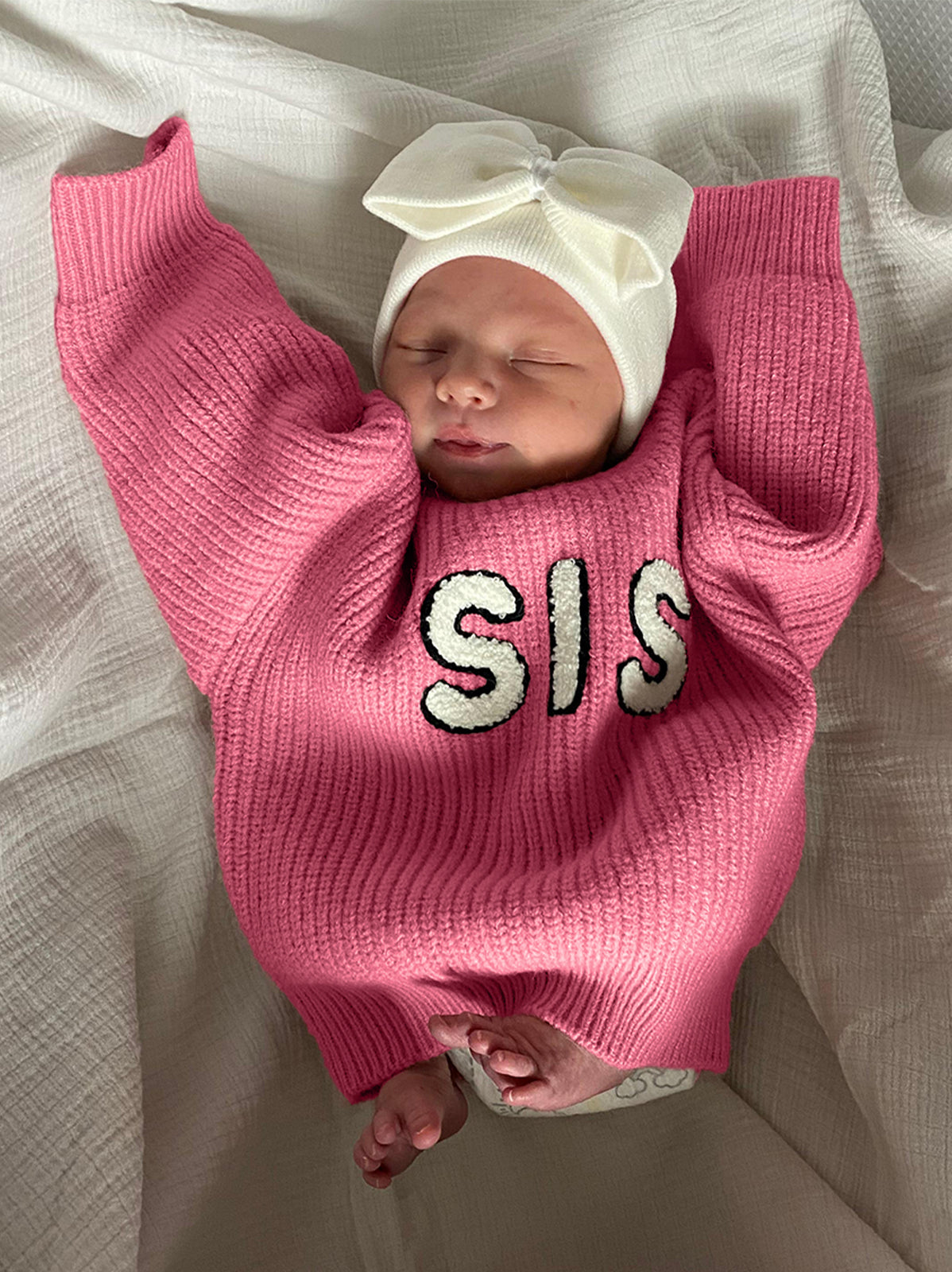 Newborn baby in a pink sweater with "SIS" text and a white bow headband, peacefully resting on a white blanket.