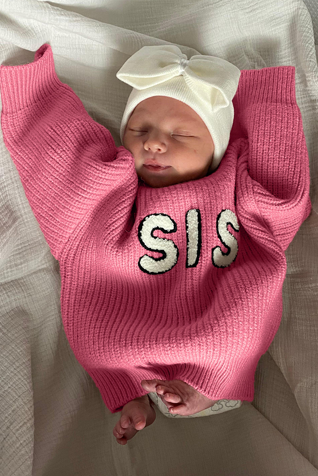 Newborn baby in a pink sweater with "SIS" text and a white bow headband, peacefully resting on a white blanket.