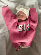 Newborn baby in a pink sweater with "SIS" text and a white bow headband, peacefully resting on a white blanket.