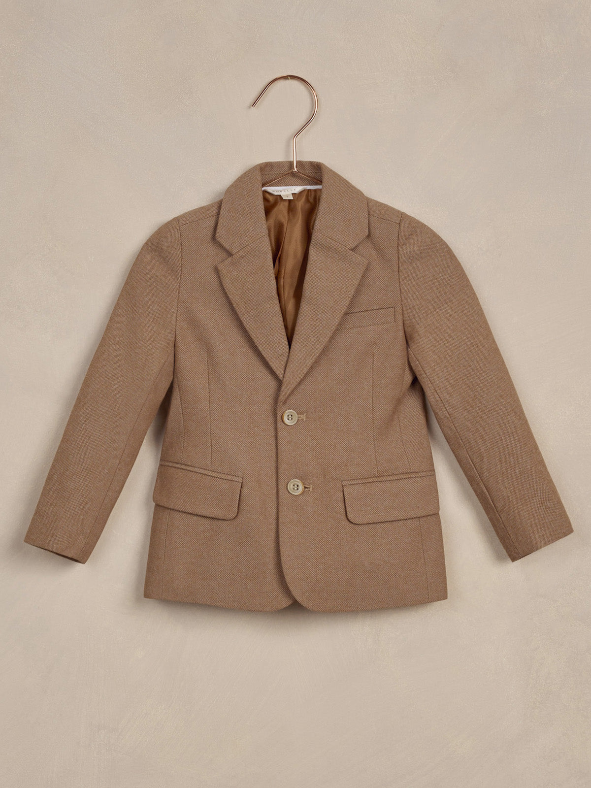 Brown child-sized blazer with notched lapels, two buttons, and front pockets, hanging on a hook.
