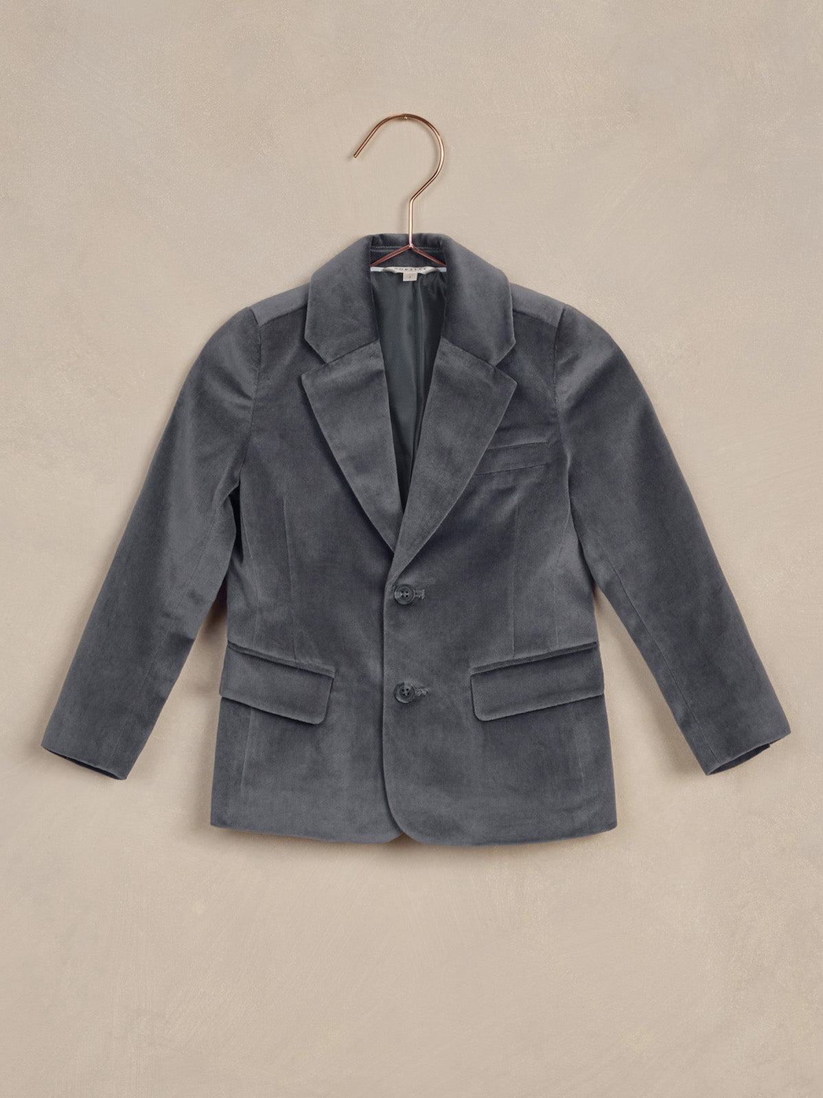 Gray velvet blazer hung on a copper hanger against a textured beige background.