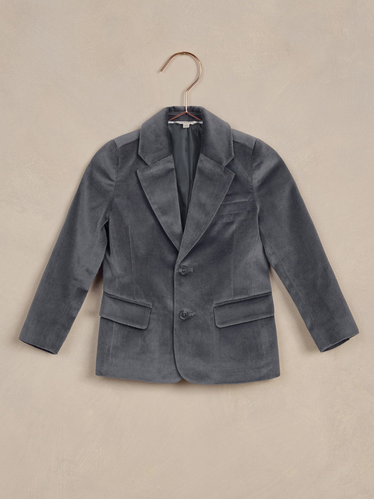Gray velvet blazer hung on a copper hanger against a textured beige background.