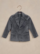 Gray velvet blazer hung on a copper hanger against a textured beige background.