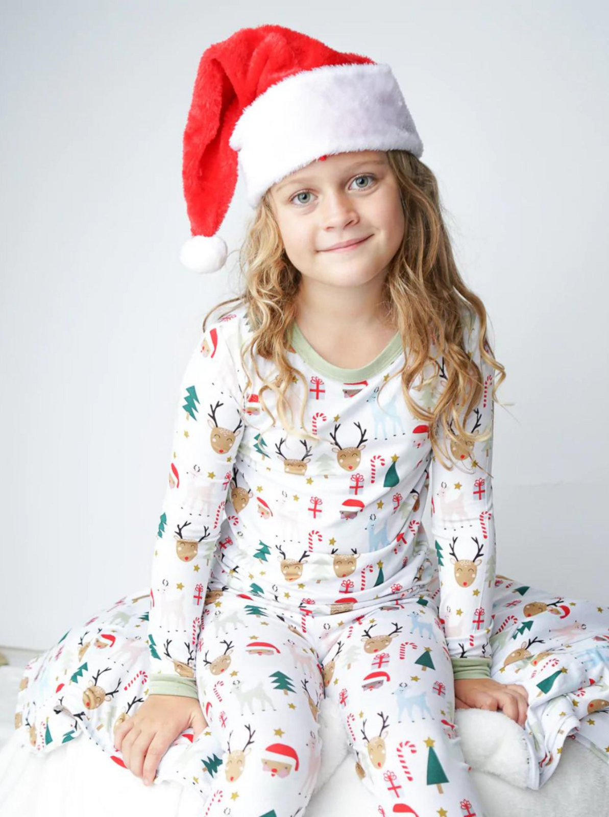 Child wearing festive pajamas and a red Santa hat, sitting on a comfortable white surface, smiling softly.