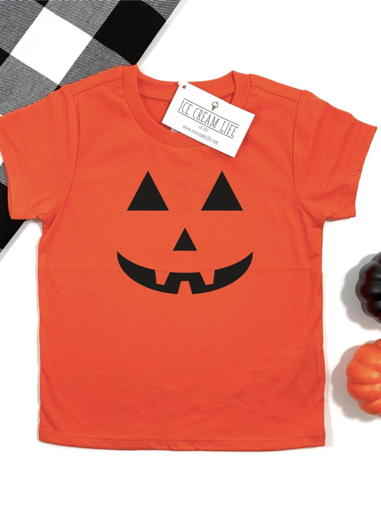 Orange Halloween shirt with black pumpkin face graphic, surrounded by black and orange pumpkins on a checkered background.