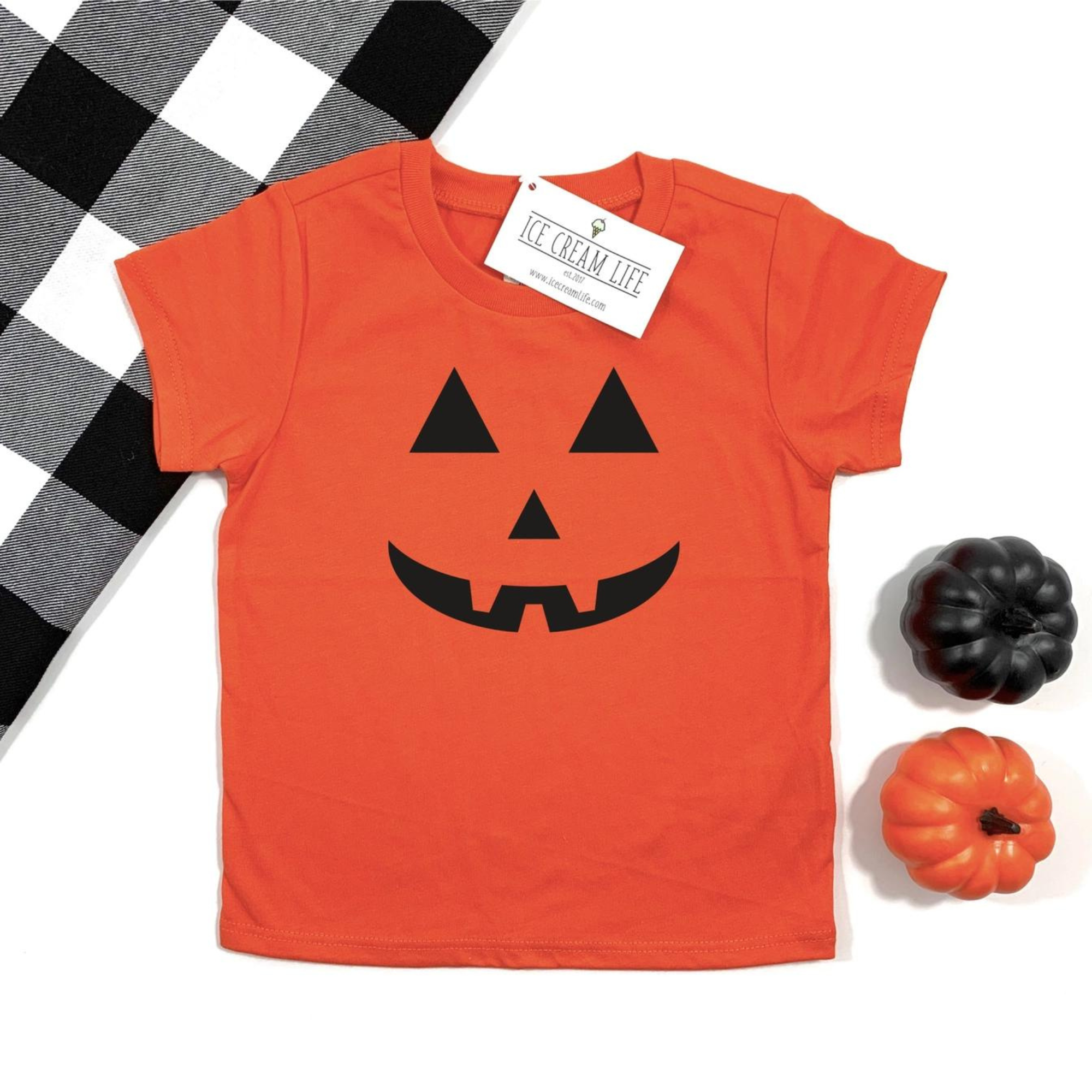 Orange Halloween shirt with black pumpkin face graphic, surrounded by black and orange pumpkins on a checkered background.
