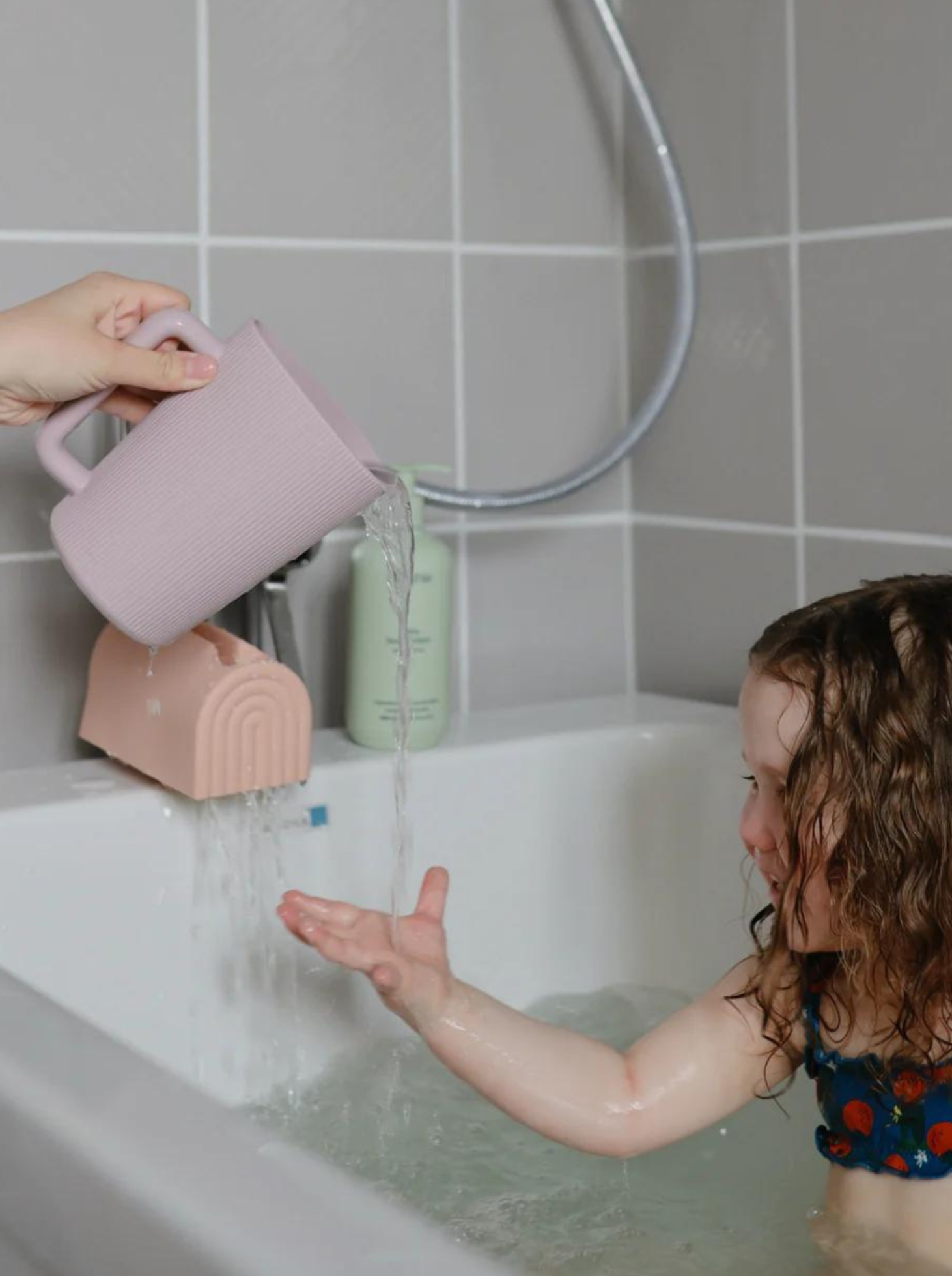 Child in a bathtub reaches out as water pours from a pink pitcher. Soft bathroom ambiance with tiled walls.