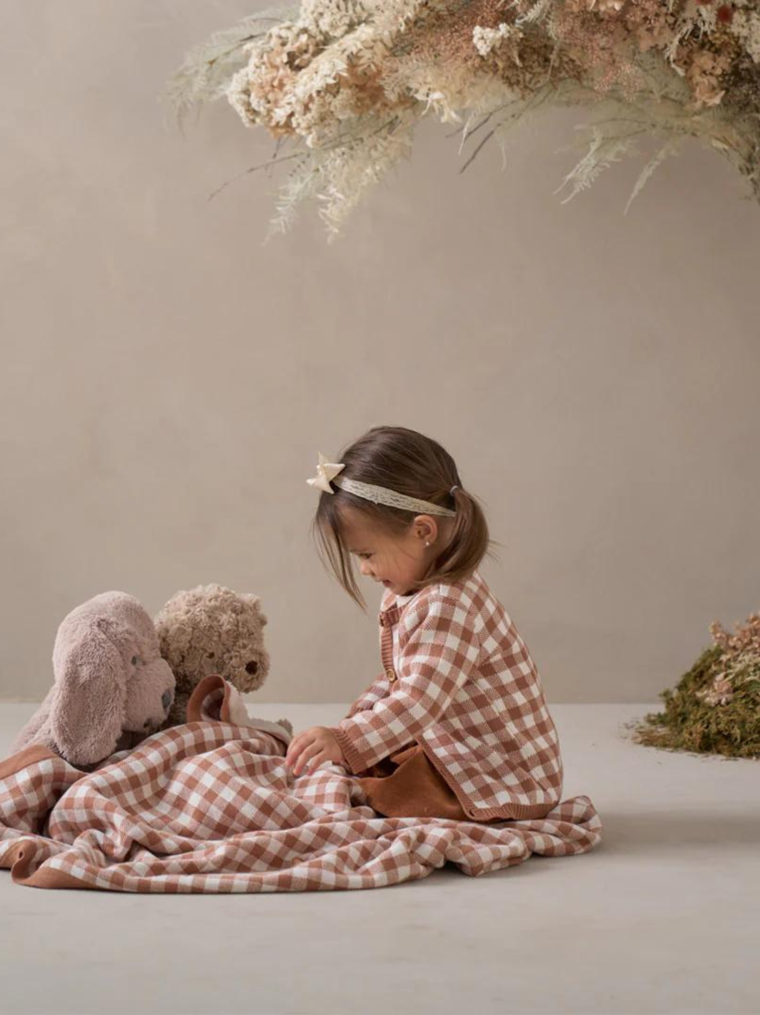 Young girl in a checkered shirt sits on a blanket with plush toys, surrounded by a serene, natural backdrop.