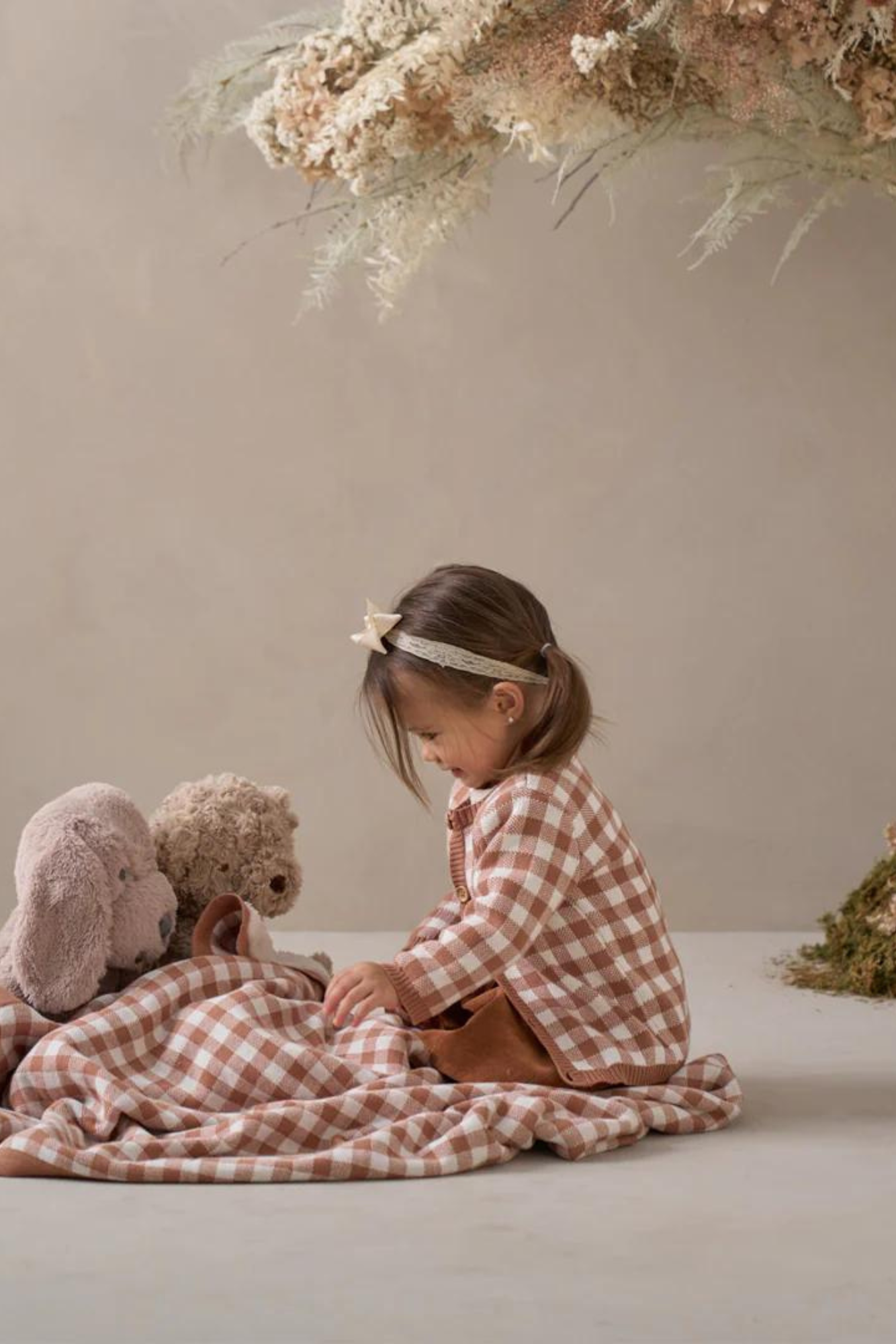 Young girl in a checkered shirt sits on a blanket with plush toys, surrounded by a serene, natural backdrop.