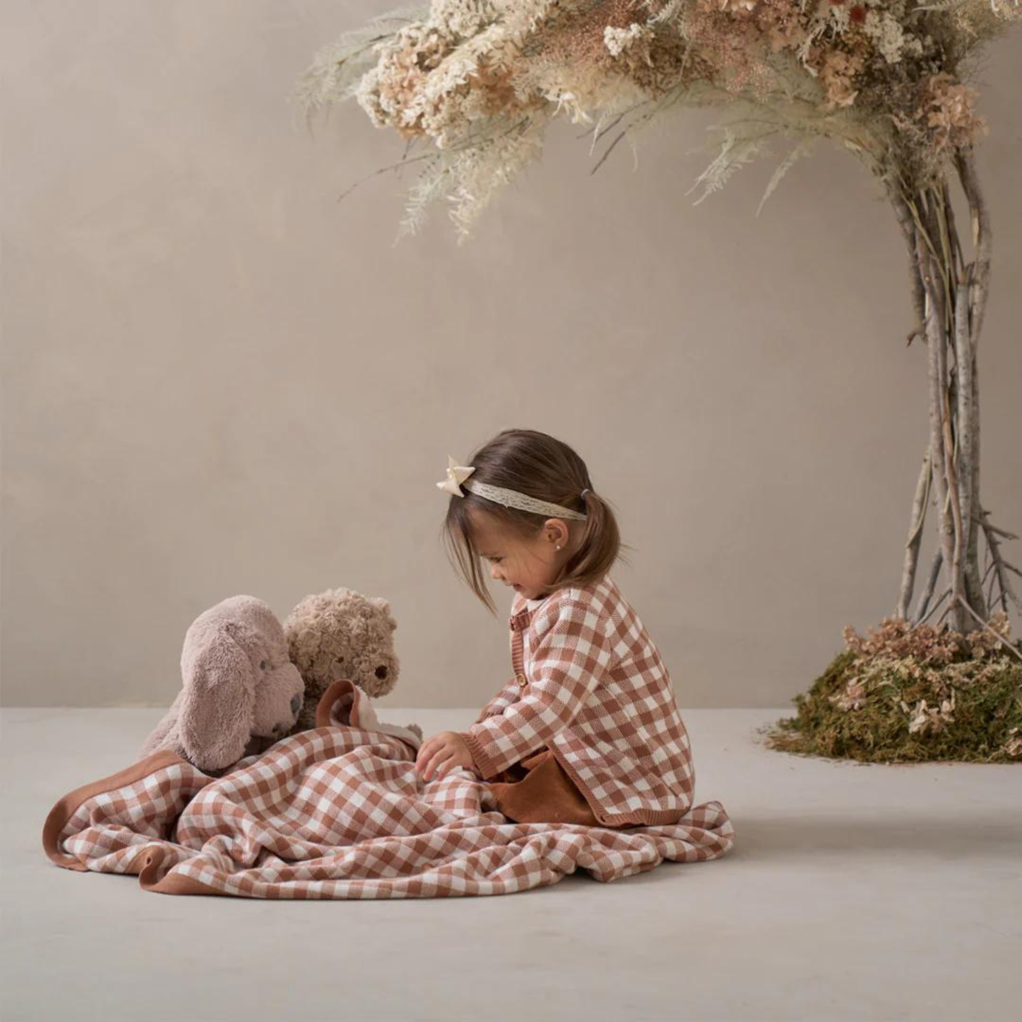 Young girl in a checkered shirt sits on a blanket with plush toys, surrounded by a serene, natural backdrop.