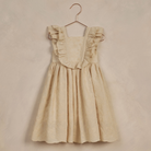 Light beige children's dress with ruffled straps hanging on a copper hanger against a textured background.