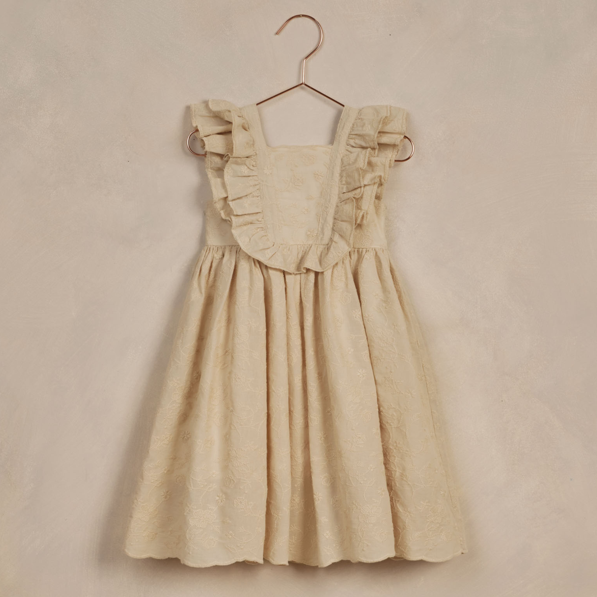 Light beige children's dress with ruffled straps hanging on a copper hanger against a textured background.