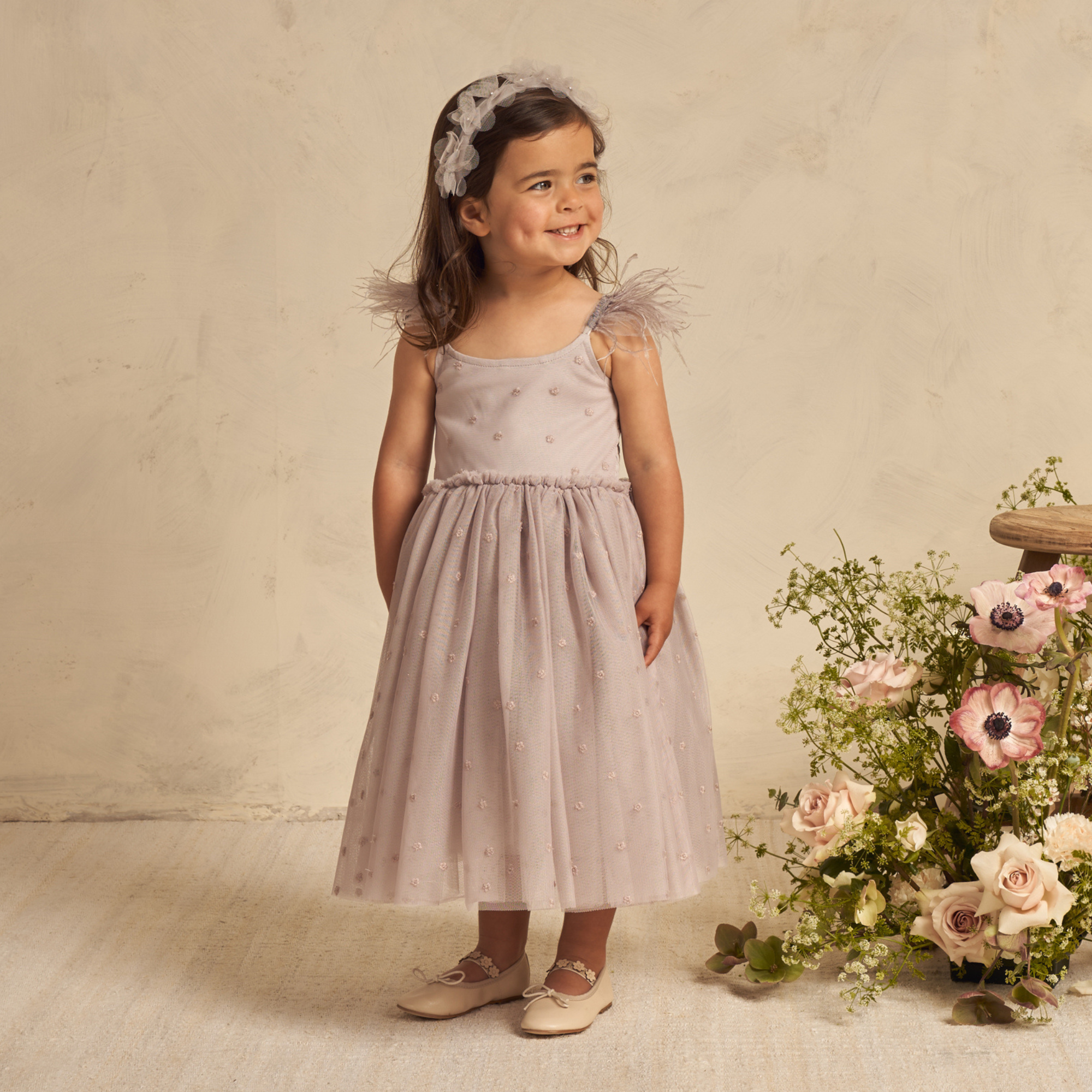 Smiling girl in a lavender dress with feathered straps, surrounded by flowers, standing on a light backdrop.