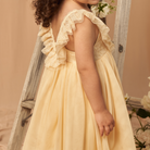 Child in a yellow dress with lace ruffles, standing next to a wooden ladder and floral backdrop.