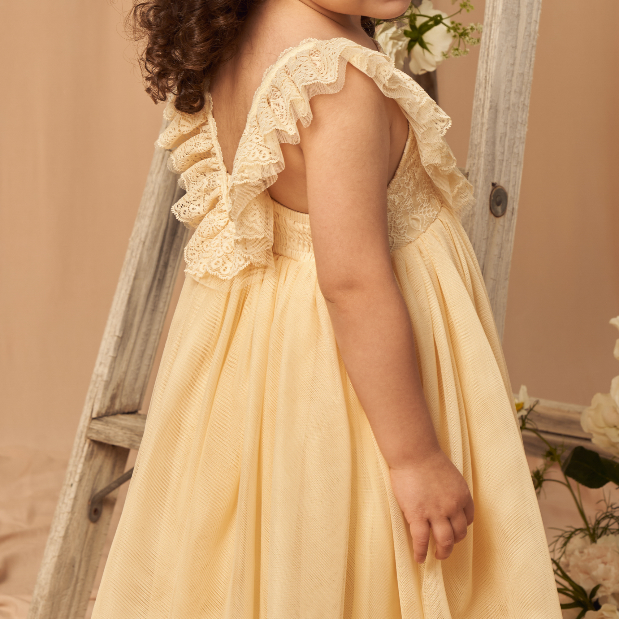Child in a yellow dress with lace ruffles, standing next to a wooden ladder and floral backdrop.