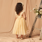 Young girl in a yellow dress with ruffled straps, standing by a wooden ladder and floral arrangement.