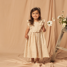 Smiling girl in cream dress holding a flower, standing on a beige backdrop next to a wooden ladder with daisies.