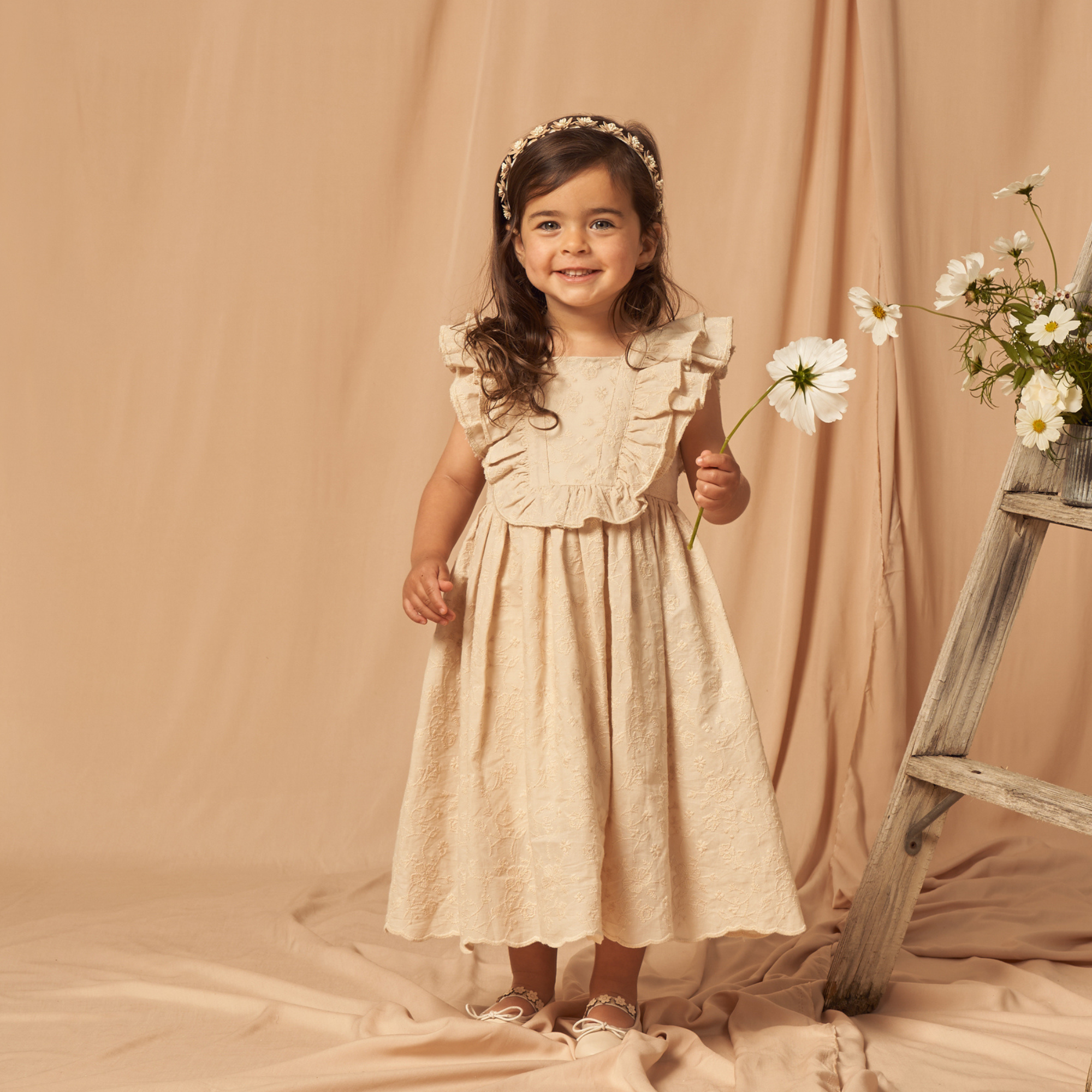 Smiling girl in cream dress holding a flower, standing on a beige backdrop next to a wooden ladder with daisies.
