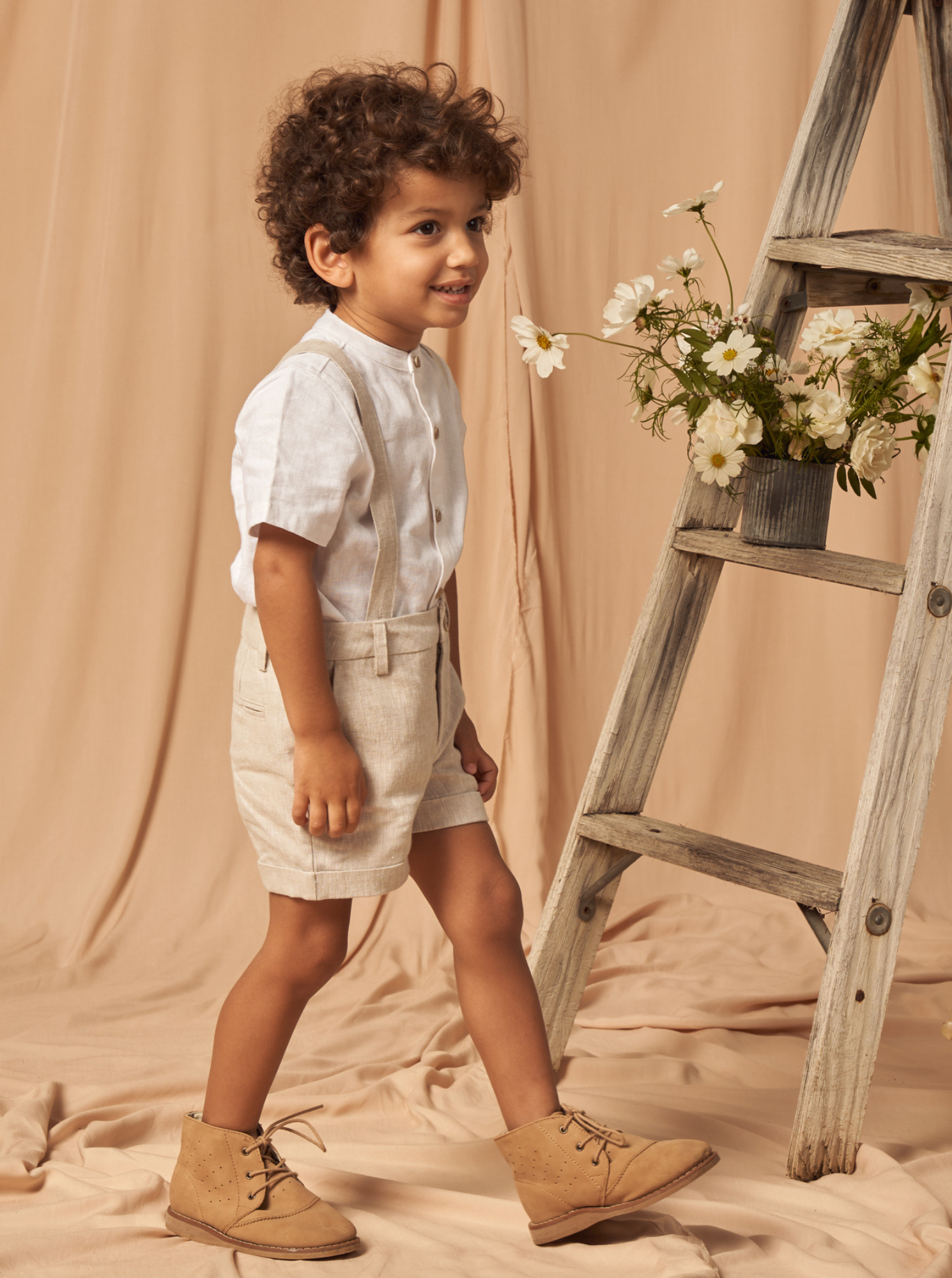 Smiling boy with curly hair in a white shirt and shorts, standing next to a wooden ladder adorned with flowers.