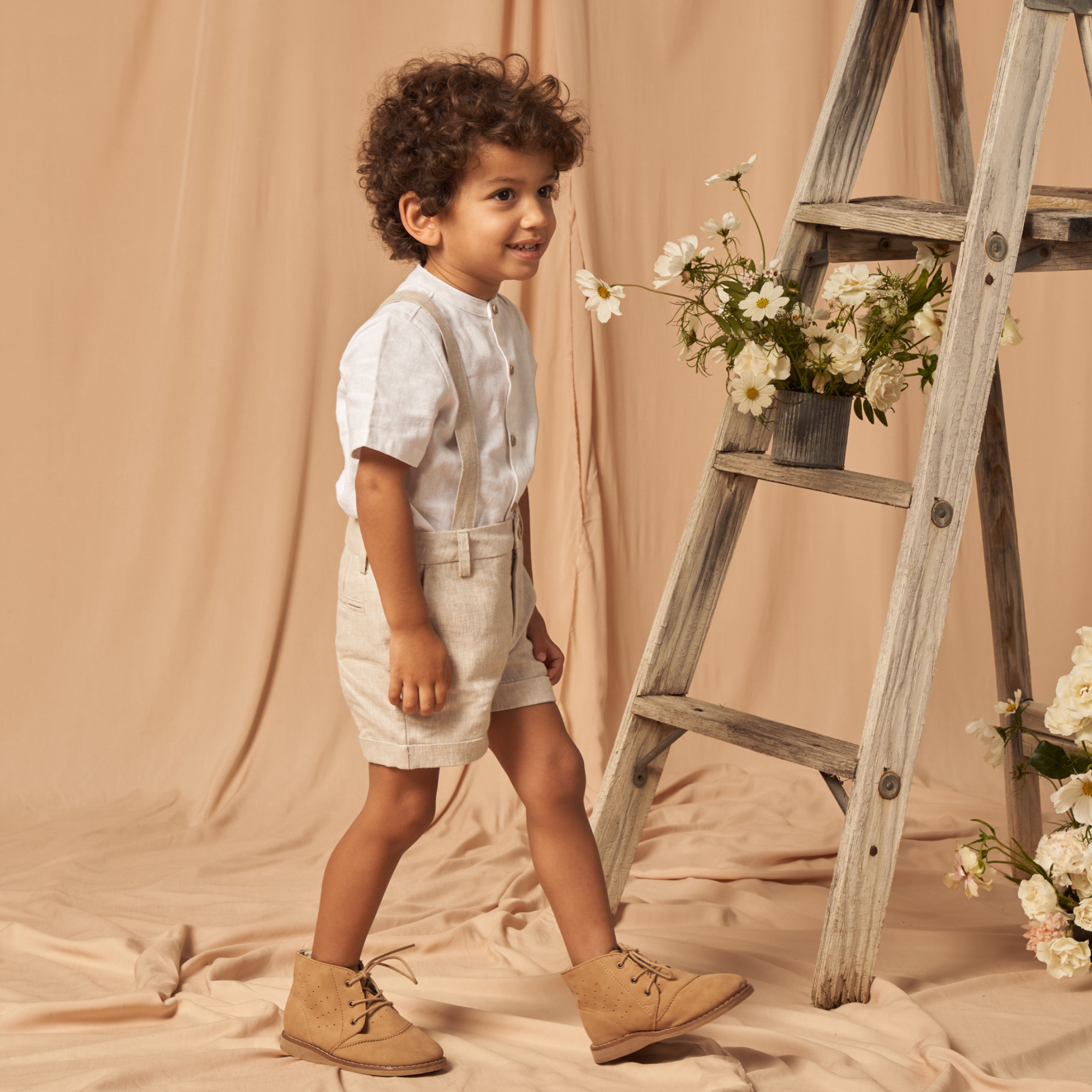Smiling boy with curly hair in a white shirt and shorts, standing next to a wooden ladder adorned with flowers.