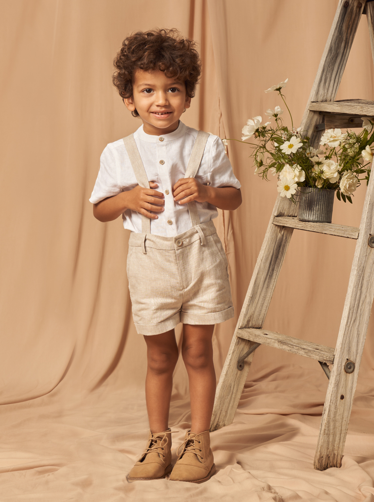 Child with curly hair in a white shirt and beige shorts stands by a wooden ladder adorned with flowers.