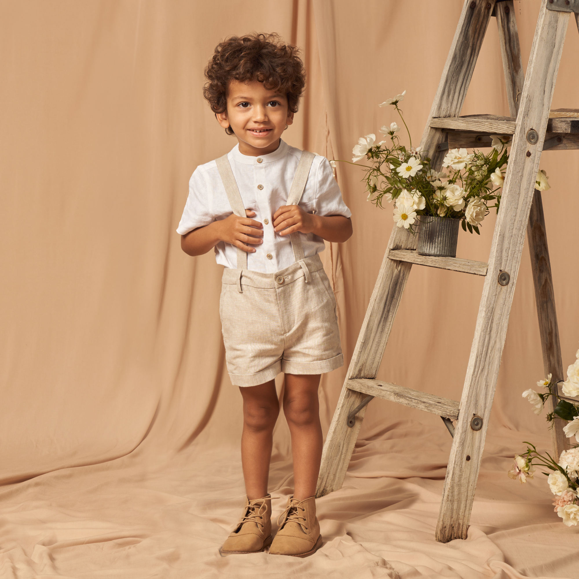 Child with curly hair in a white shirt and beige shorts stands by a wooden ladder adorned with flowers.