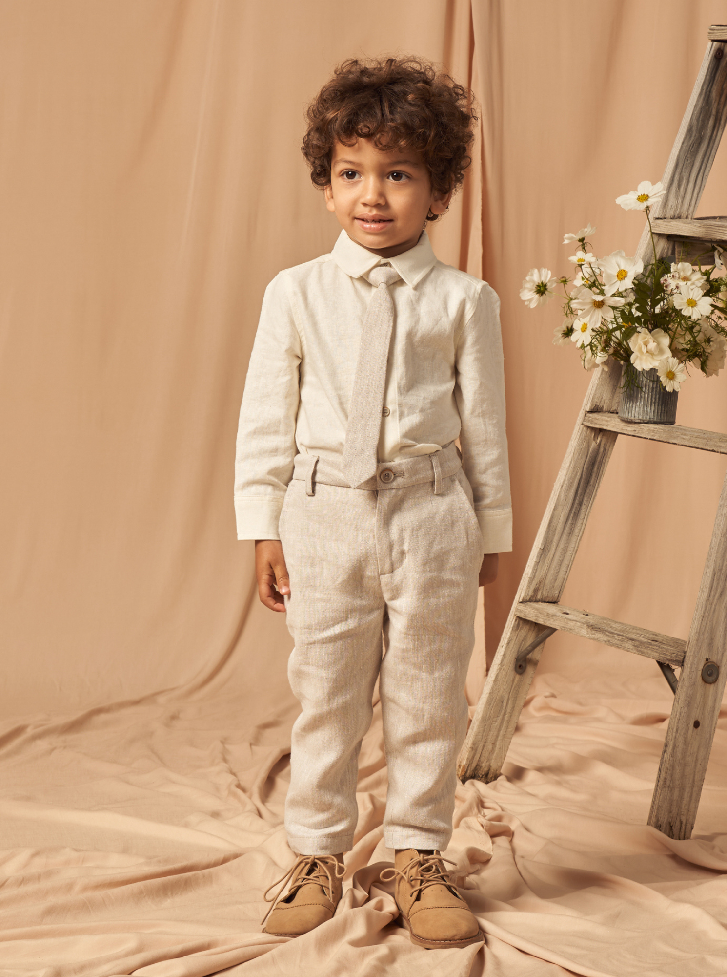 Young boy in a cream suit with a tie, standing beside a decorative ladder and flowers against a beige backdrop.