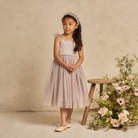 Girl in a lavender dress poses by a wooden stool adorned with flowers against a soft beige backdrop.