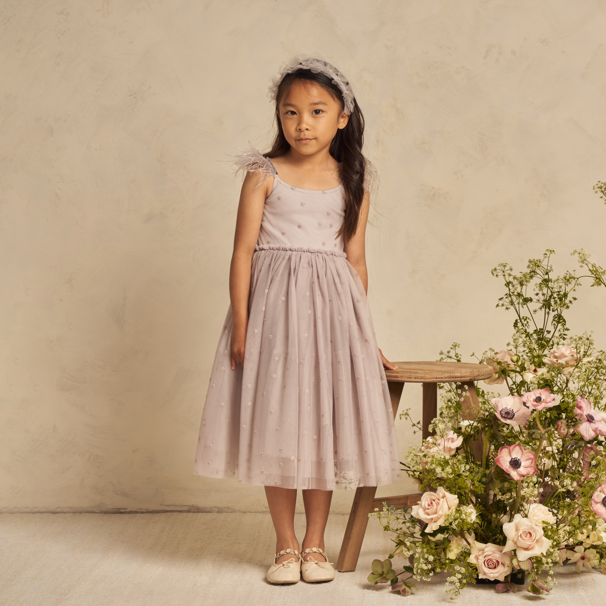 Girl in a lavender dress with floral details stands beside a wooden stool and a bouquet of pastel flowers.