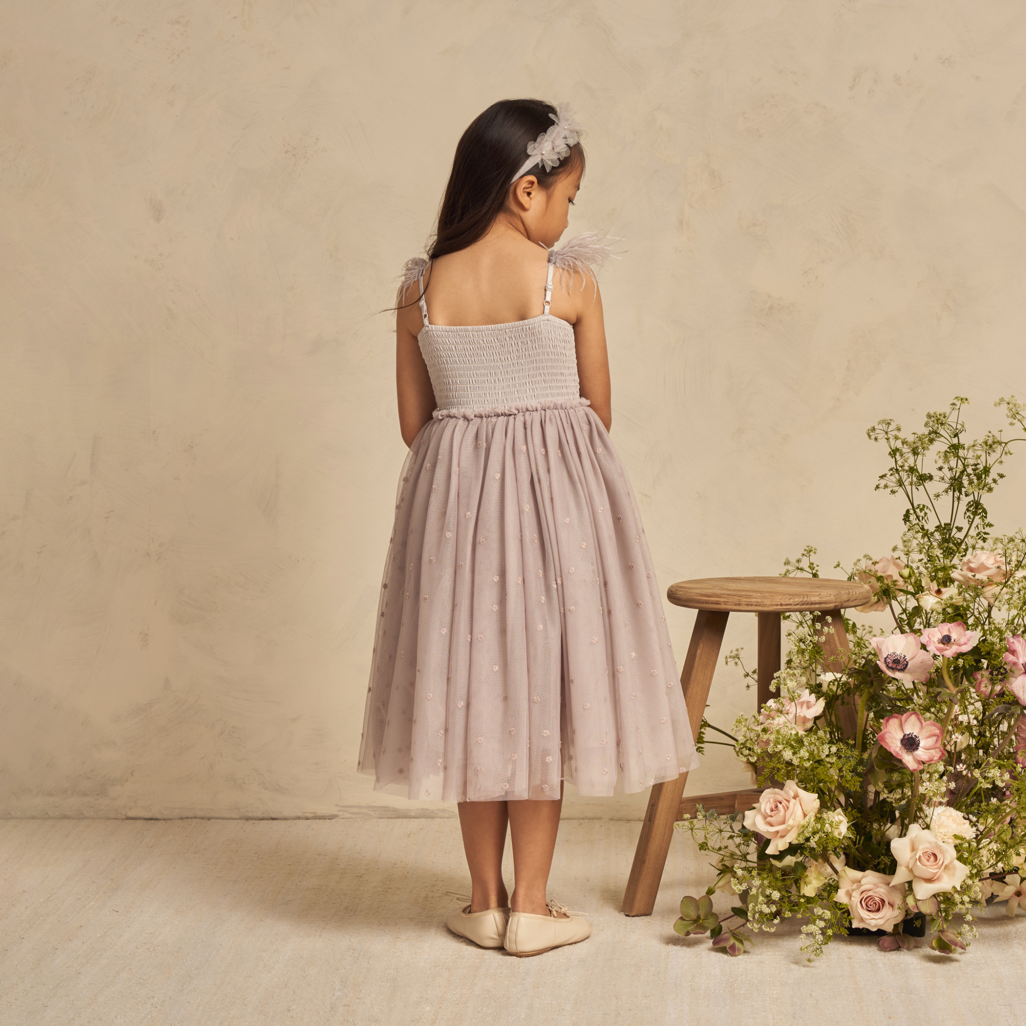 Girl in a floral dress with a smocked bodice, standing by a wooden stool decorated with fresh flowers.
