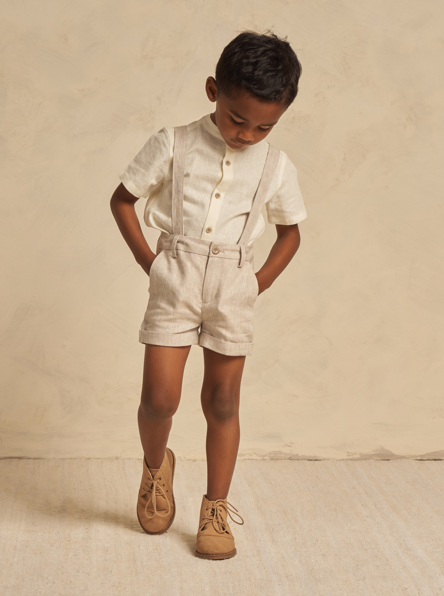 Child in light-colored outfit with suspenders, shorts, and brown shoes, looking down against a textured beige background.