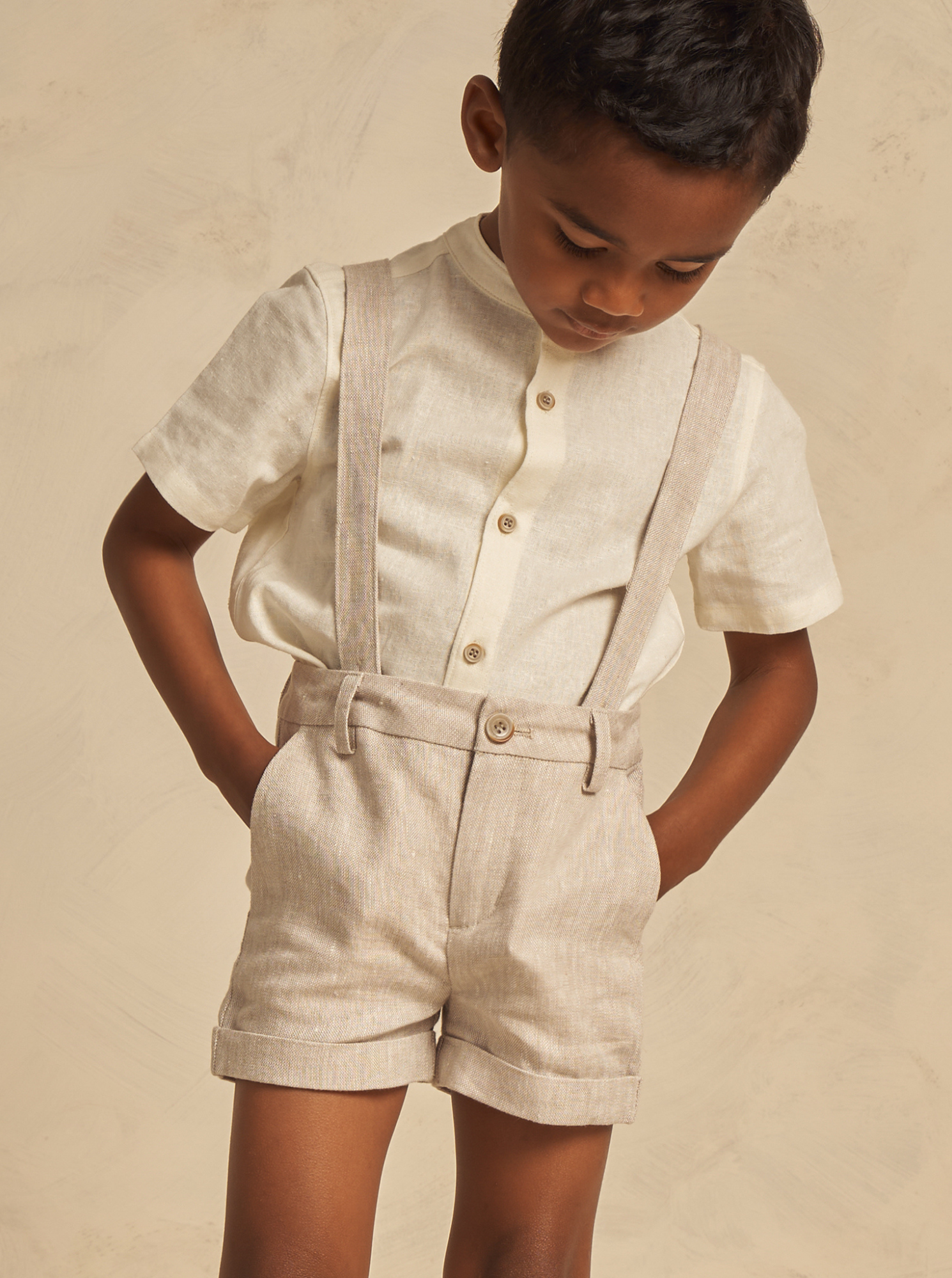 Child wearing a light beige shirt and shorts with suspenders, looking down against a neutral background.