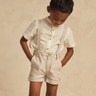 Child wearing a light beige shirt and shorts with suspenders, looking down against a neutral background.